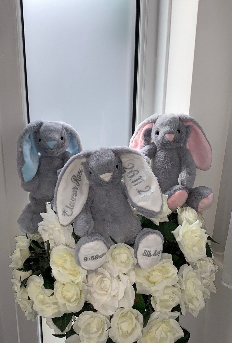May include: Three gray plush bunny rabbits with embroidered details. The middle bunny has the text "Eleanor Rose" and "26.11.22" on its ears. The bunny on the left has blue ears and the bunny on the right has pink ears. All three bunnies are sitting in a bouquet of white roses.
