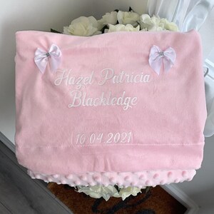 May include: A pink baby blanket with white embroidered text that reads "Hazel Patricia Blackledge 10.04.2021". The blanket has two white bows with silver embellishments.