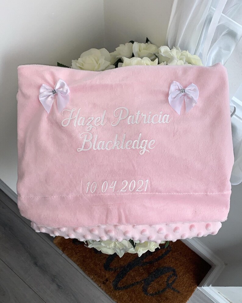 May include: A pink baby blanket with white embroidered text that reads "Hazel Patricia Blackledge 10.04.2021". The blanket has two white bows with silver embellishments.