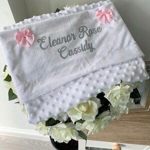 May include: White baby blanket with a soft, textured surface. The blanket has a gray embroidered name "Eleanor Rose Cassidy" and two pink bows.