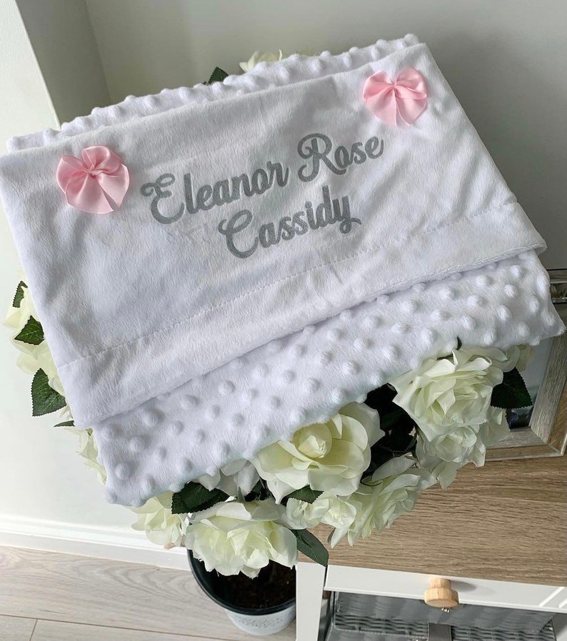 May include: White baby blanket with a soft, textured surface. The blanket has a gray embroidered name "Eleanor Rose Cassidy" and two pink bows.