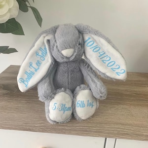May include: A gray plush bunny with white ears. The ears are embroidered with the text "Bodhi Lee Sellers" and "10/04/2022". The bunny's feet are embroidered with "5:31pm" and "6lb 1oz".