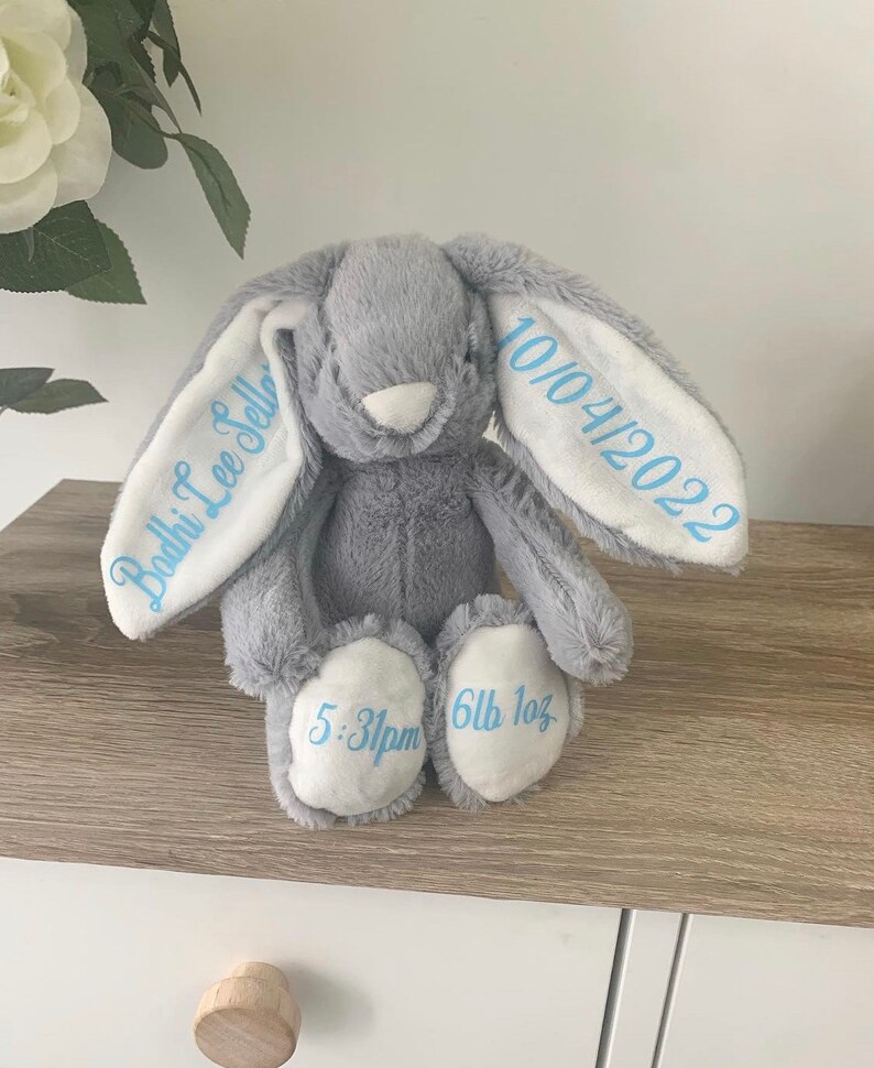 May include: A gray plush bunny with white ears. The ears are embroidered with the text "Bodhi Lee Sellers" and "10/04/2022". The bunny's feet are embroidered with "5:31pm" and "6lb 1oz".