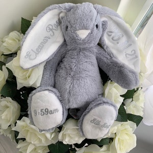 May include: A gray plush bunny with white ears. The ears are embroidered with the text "Eleanor Rose" and "05.11.22". The bunny's paws are embroidered with "9:59am" and "Little Sof"