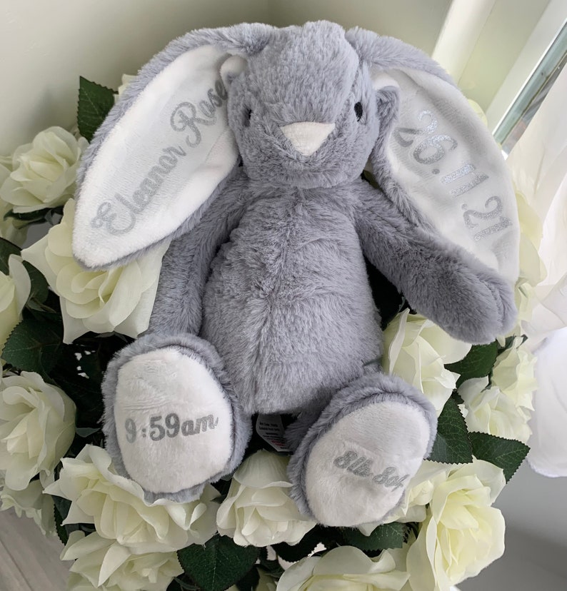 May include: A gray plush bunny with white ears. The ears are embroidered with the text "Eleanor Rose" and "05.11.22". The bunny's paws are embroidered with "9:59am" and "Little Sof"