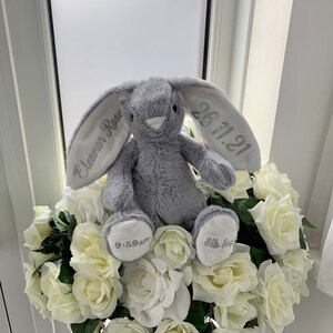 May include: A gray plush bunny rabbit with embroidered ears. The left ear says "Eleanor Rose" and the right ear says "26.11.21". The bunny is sitting on a bed of white roses.