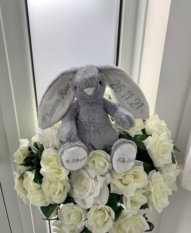 May include: A gray plush bunny rabbit with embroidered ears. The left ear says "Eleanor Rose" and the right ear says "26.11.21". The bunny is sitting on a bed of white roses.