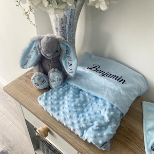 May include: A light blue baby blanket with a gray bunny plush toy. The blanket has a textured surface and is embroidered with the name "Benjamin" and the date "07.02.2023". The bunny toy is embroidered with "Benjamin Peter Graham" and the baby's birth weight and time.