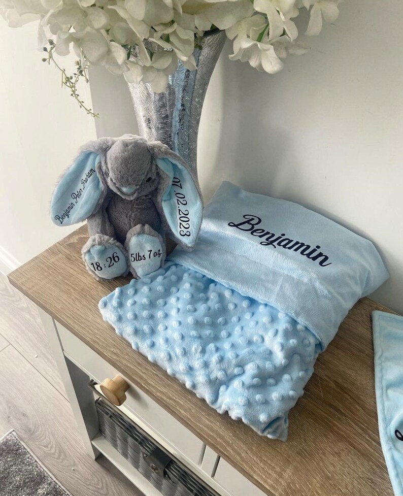 May include: A light blue baby blanket with a gray bunny plush toy. The blanket has a textured surface and is embroidered with the name "Benjamin" and the date "07.02.2023". The bunny toy is embroidered with "Benjamin Peter Graham" and the baby's birth weight and time.
