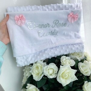 May include: White baby blanket with a silver embroidered name "Eleanor Rose Cassidy" and two pink bows.