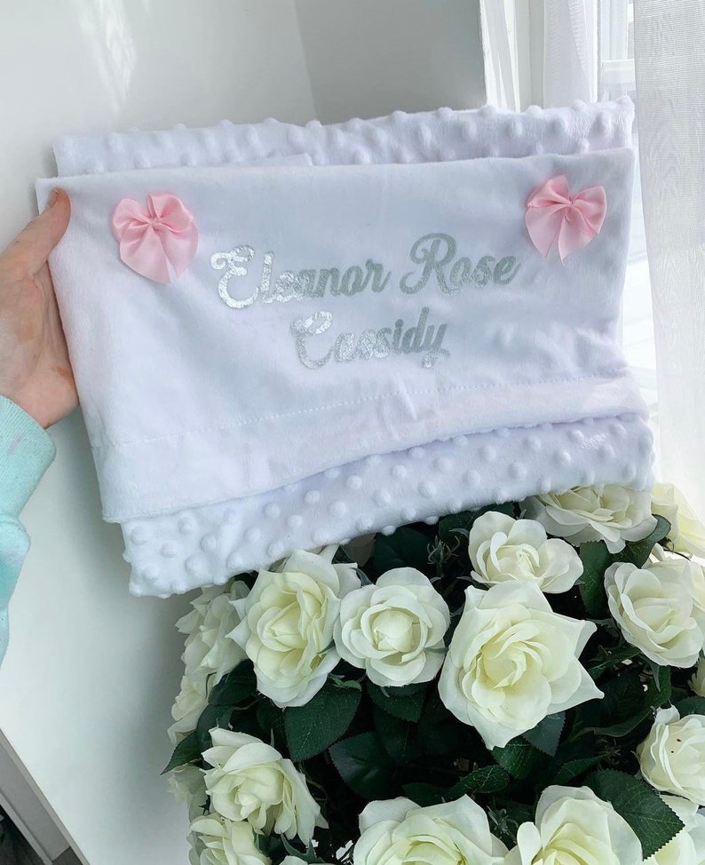 May include: White baby blanket with a silver embroidered name "Eleanor Rose Cassidy" and two pink bows.