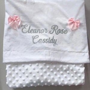 May include: White baby blanket with a soft, textured surface. The blanket has a silver glitter embroidered name "Eleanor Rose Cassidy" and two pink satin bows.