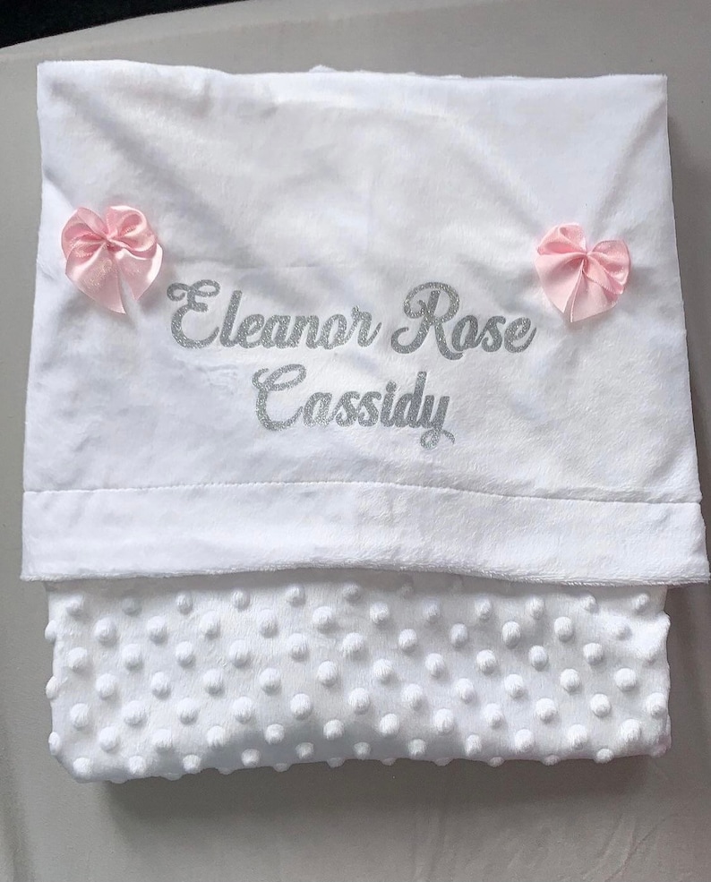 May include: White baby blanket with a soft, textured surface. The blanket has a silver glitter embroidered name "Eleanor Rose Cassidy" and two pink satin bows.