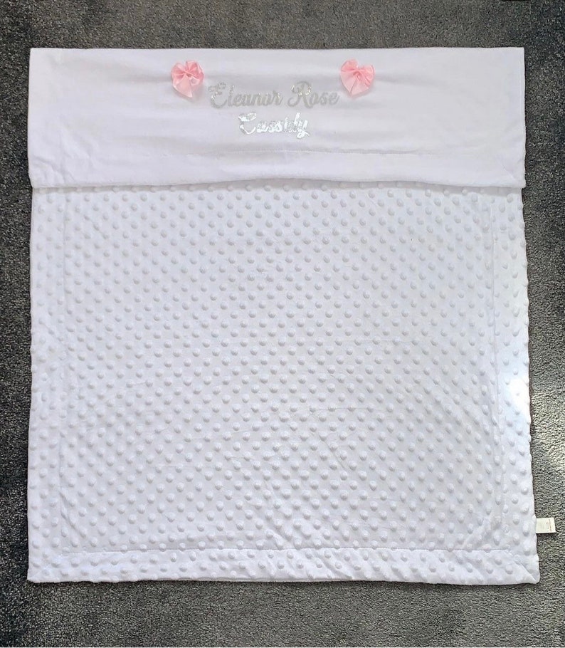 May include: White baby blanket with a white satin border and pink satin bows. The blanket is personalized with the name "Eleanor Rose" and the word "Sweetly" in silver embroidery.