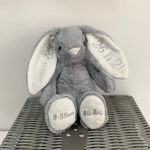May include: A gray plush bunny with white ears. The ears are embroidered with the text "26.11.21" and "Eleanor Rose". The bunny's paws are embroidered with "9:59am" and "8lb 8oz".