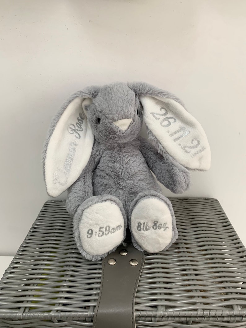 May include: A gray plush bunny with white ears. The ears are embroidered with the text "26.11.21" and "Eleanor Rose". The bunny's paws are embroidered with "9:59am" and "8lb 8oz".