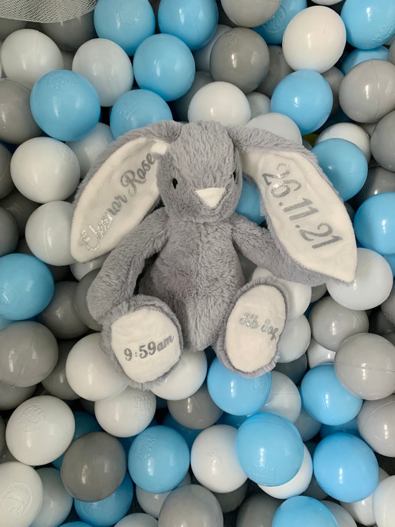 May include: A gray plush bunny with embroidered ears. The left ear says "Eleanor Rose" and the right ear says "26.11.21". The bunny is sitting in a ball pit filled with blue, white, and gray balls. The bunny's foot says "9:59am".