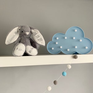 May include: A gray bunny plush toy with embroidered text on its ears, "Cameron Rose" and "26.11.21". The bunny is sitting on a white shelf with a blue cloud-shaped night light with white LED lights. A white string with blue, gray, and white pom-poms hangs from the shelf.