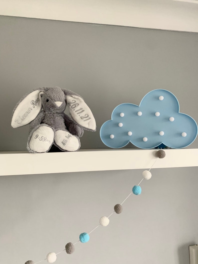 May include: A gray bunny plush toy with embroidered text on its ears, "Cameron Rose" and "26.11.21". The bunny is sitting on a white shelf with a blue cloud-shaped night light with white LED lights. A white string with blue, gray, and white pom-poms hangs from the shelf.