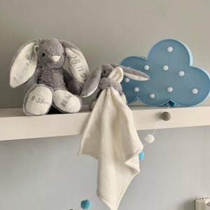 May include: Two gray plush bunny toys with embroidered details, one with the text "Eleanor Rose" and "26.11.21" and the other with "Ella Joy". A blue cloud-shaped nightlight with white lights is on a white shelf with a white and blue pom-pom garland.