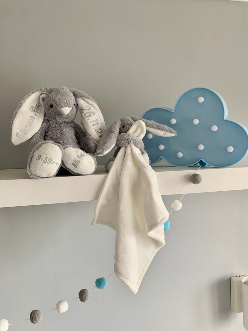 May include: Two gray plush bunny toys with embroidered details, one with the text "Eleanor Rose" and "26.11.21" and the other with "Ella Joy". A blue cloud-shaped nightlight with white lights is on a white shelf with a white and blue pom-pom garland.