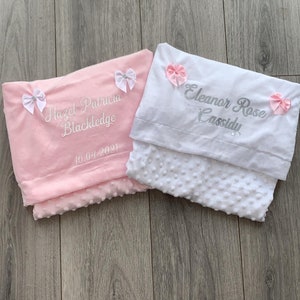 May include: Two personalized baby blankets, one pink and one white, with embroidered names and dates. The pink blanket has the name "Hazel Patricia Blackledge" and the date "10.04.2021". The white blanket has the name "Eleanor Rose Cassidy". Both blankets have pink bows.