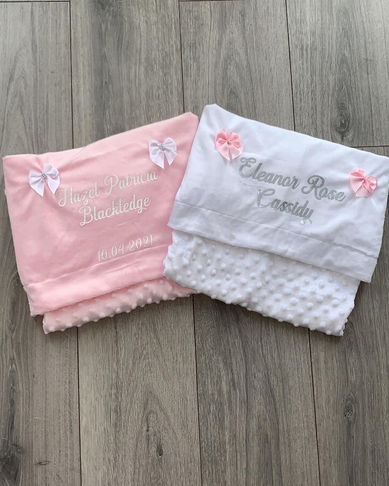 May include: Two personalized baby blankets, one pink and one white, with embroidered names and dates. The pink blanket has the name "Hazel Patricia Blackledge" and the date "10.04.2021". The white blanket has the name "Eleanor Rose Cassidy". Both blankets have pink bows.
