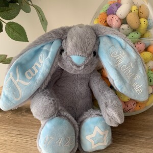 May include: A gray plush bunny with blue ears and feet. The ears are embroidered with "My First Easter" and "Kane". The feet are embroidered with "17.4.22" and a white star.