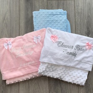 May include: Two personalized baby blankets, one pink and one white. The pink blanket has the name "Hazel Patricia Blackledge" and the date "10.04.2021" embroidered on it. The white blanket has the name "Eleanor Rose Cassidy" embroidered on it.