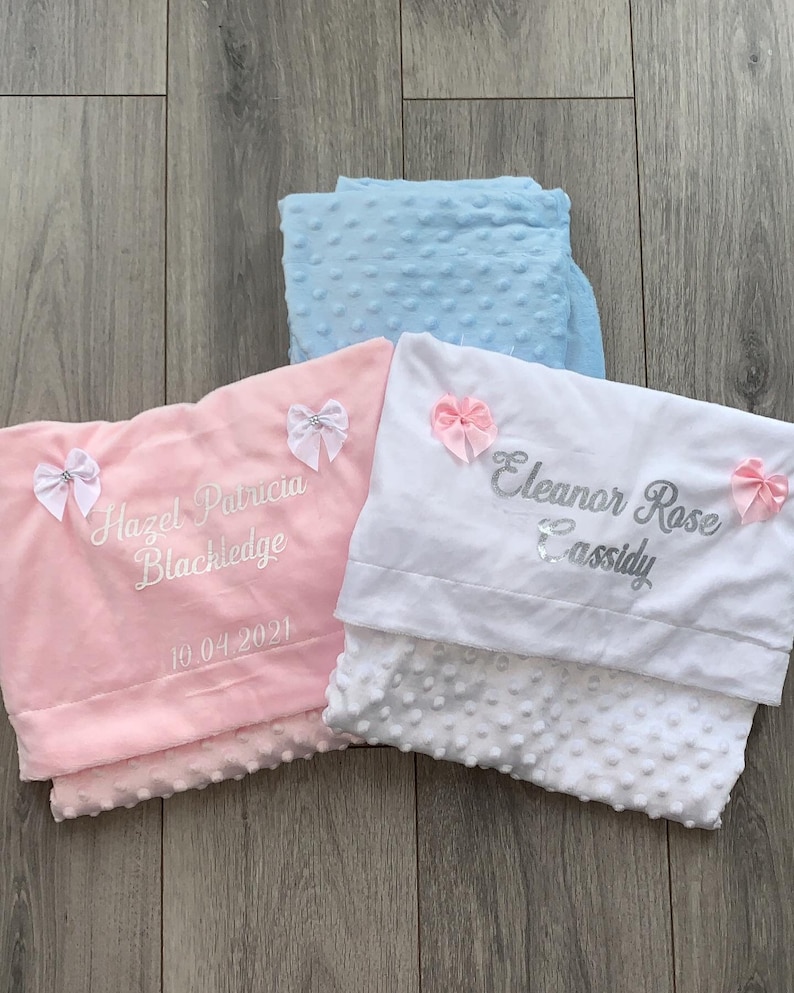 May include: Two personalized baby blankets, one pink and one white. The pink blanket has the name "Hazel Patricia Blackledge" and the date "10.04.2021" embroidered on it. The white blanket has the name "Eleanor Rose Cassidy" embroidered on it.