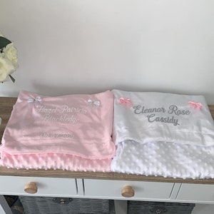 May include: Two personalized baby blankets, one pink and one white. The pink blanket has the text "Hazel Patricia Blackledge 10.04.2021" embroidered on it. The white blanket has the text "Eleanor Rose Cassidy" embroidered on it.