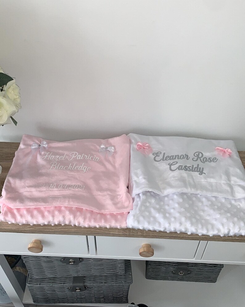 May include: Two personalized baby blankets, one pink and one white. The pink blanket has the text "Hazel Patricia Blackledge 10.04.2021" embroidered on it. The white blanket has the text "Eleanor Rose Cassidy" embroidered on it.