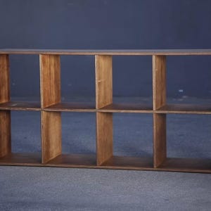 May include: A rectangular wooden shelf unit with eight open compartments. The unit is constructed from light brown wood and has a simple, modern design. The shelves are evenly spaced, creating a grid-like appearance. The unit is suitable for storage or display.