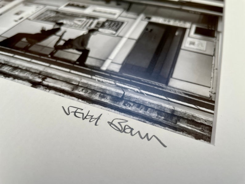 May include: Black and white photograph of a street scene, featuring architectural details and figures. The image is signed in the lower left corner. The composition includes a building facade, a door, and people.
