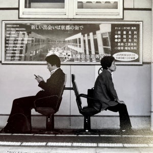 May include: Black and white photograph of two people seated on chairs in a public space. One person is using a smartphone, while the other is looking away. A sign with Japanese text is visible in the background.