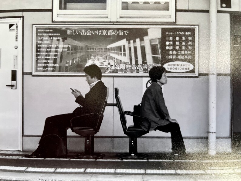 May include: Black and white photograph of two people seated on chairs in a public space. One person is using a smartphone, while the other is looking away. A sign with Japanese text is visible in the background.