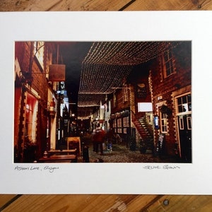 May include: A photograph of a narrow, brick-lined alleyway in Glasgow, Scotland. The alleyway is decorated with string lights and has a sign that reads "Ashton Lane, Glasgow".