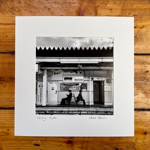 May include: Black and white photograph of a train station scene in Kyoto, Japan. Two people are seated on a bench, waiting. The image is framed with a white mat and the title "Waiting; Kyoto" is written at the bottom.