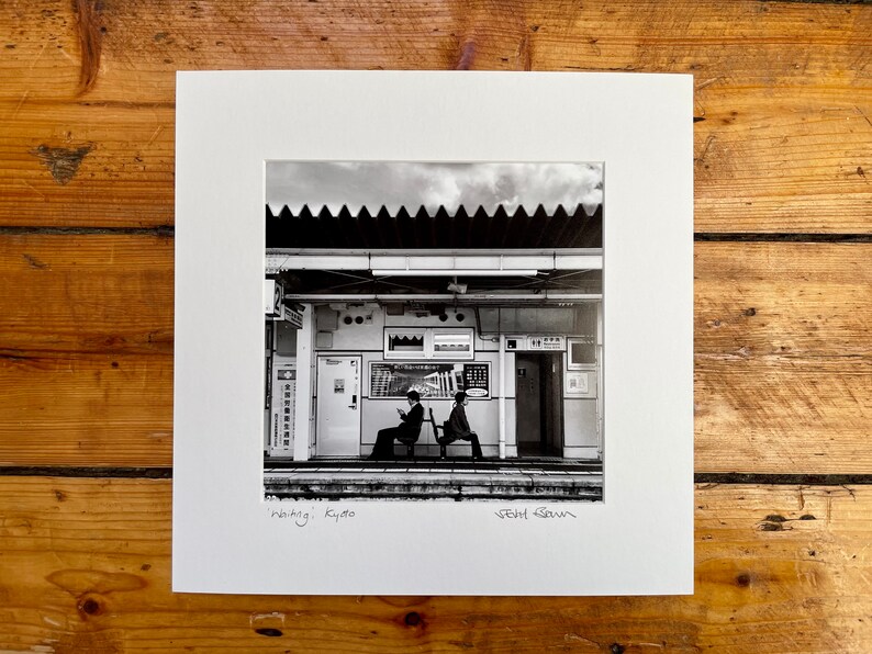 May include: Black and white photograph of a train station scene in Kyoto, Japan. Two people are seated on a bench, waiting. The image is framed with a white mat and the title "Waiting; Kyoto" is written at the bottom.
