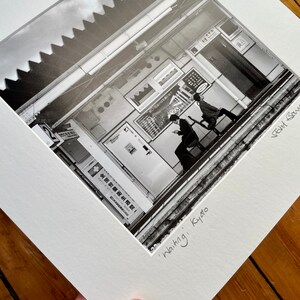 May include: Black and white photograph of a street scene in Kyoto, Japan, titled "Waiting." The image shows two people walking on a platform, with signs and architectural details in the background. The photograph is matted and framed.