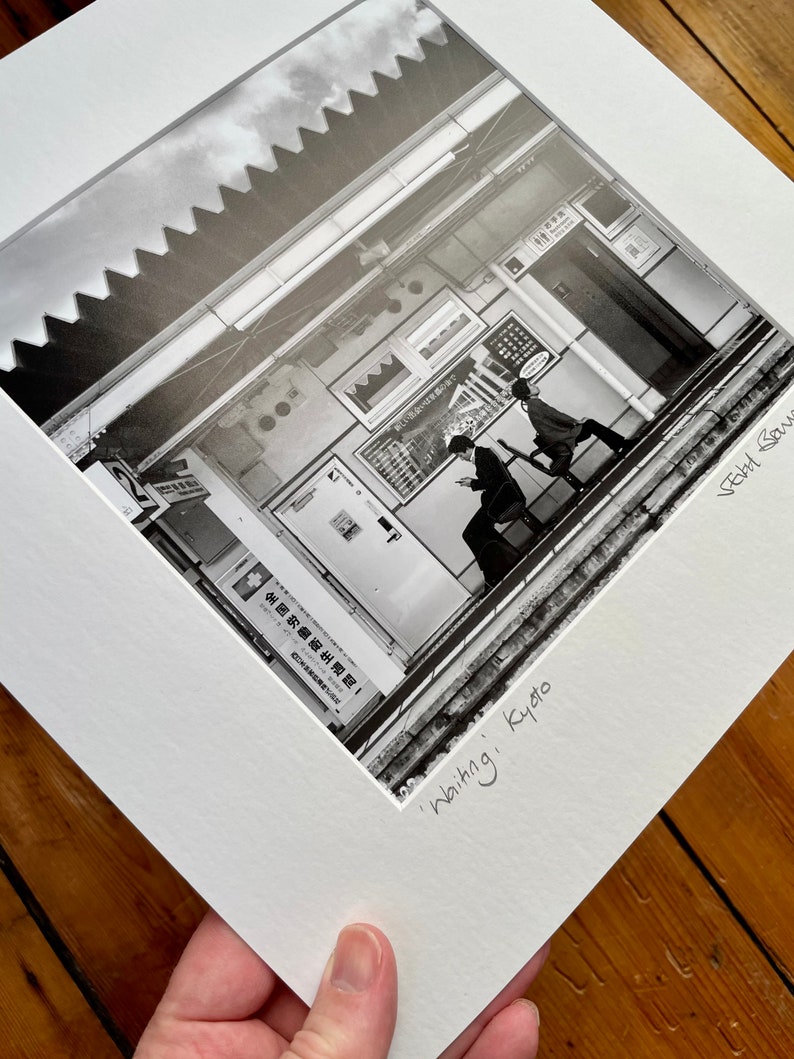 May include: Black and white photograph of a street scene in Kyoto, Japan, titled "Waiting." The image shows two people walking on a platform, with signs and architectural details in the background. The photograph is matted and framed.