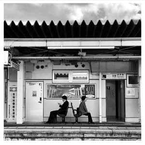 May include: Black and white photograph of a train station platform. Two people sit on metal chairs, waiting. The station has a corrugated roof and signage in Japanese. The sky is overcast.