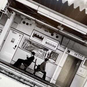 May include: Black and white photograph of a train station platform. Two people are seated on benches. Signage includes Japanese text and a restroom symbol. The station has a modern design with overhead lighting and a corrugated roof.