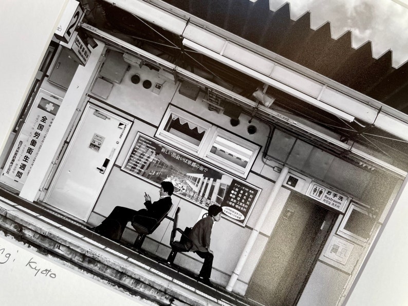 May include: Black and white photograph of a train station platform. Two people are seated on benches. Signage includes Japanese text and a restroom symbol. The station has a modern design with overhead lighting and a corrugated roof.