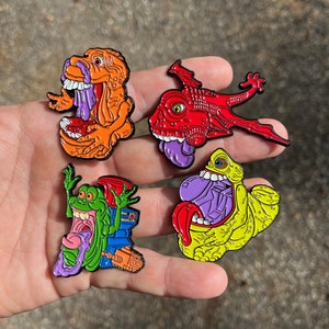 May include: Four enamel pins featuring characters from the Ghostbusters franchise. The pins are colorful and feature a variety of designs, including a green ghost, a red dragon, a yellow monster, and an orange monster.