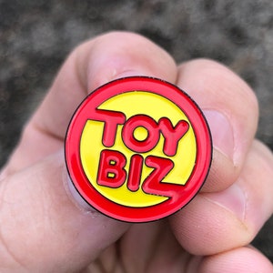 May include: A red and yellow enamel pin with the words "TOY BIZ" in a bold, white font.