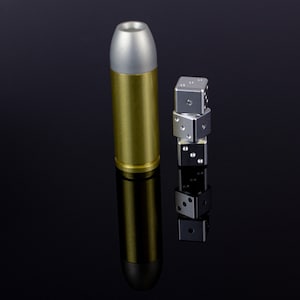 May include: A metallic gold bullet-shaped object with a silver tip stands next to a stack of four silver dice. The dice are stacked vertically, each showing a different number of dots. The objects are on a reflective black surface.