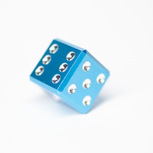 Puede incluir: Un dado de metal azul con puntos blancos. El dado est&aacute; tumbado de lado.