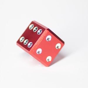 Puede incluir: Un dado de metal rojo con puntos blancos en cada lado. El dado est&aacute; acostado de lado con el n&uacute;mero cuatro hacia arriba.