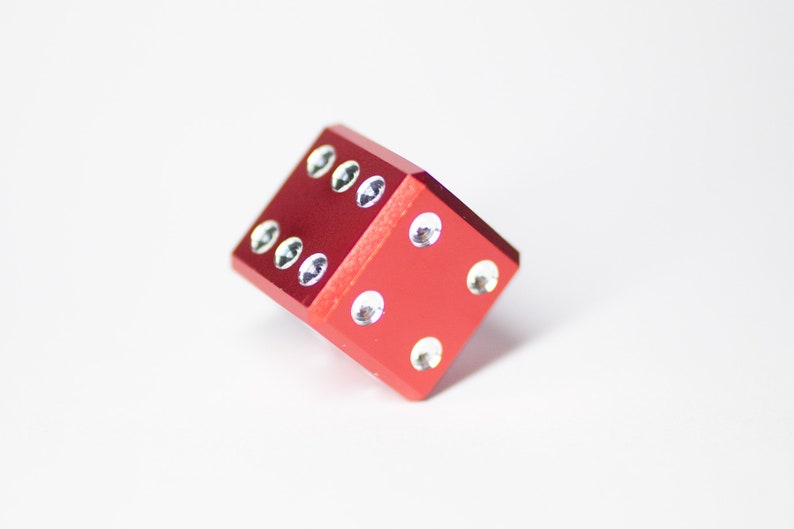 Puede incluir: Un dado de metal rojo con puntos blancos en cada lado. El dado est&aacute; acostado de lado con el n&uacute;mero cuatro hacia arriba.